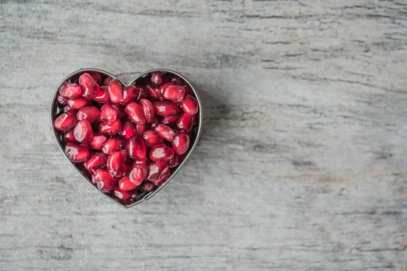 Delicious and juicy pomegranate seeds in a heart-shaped bowl on a wooden table.